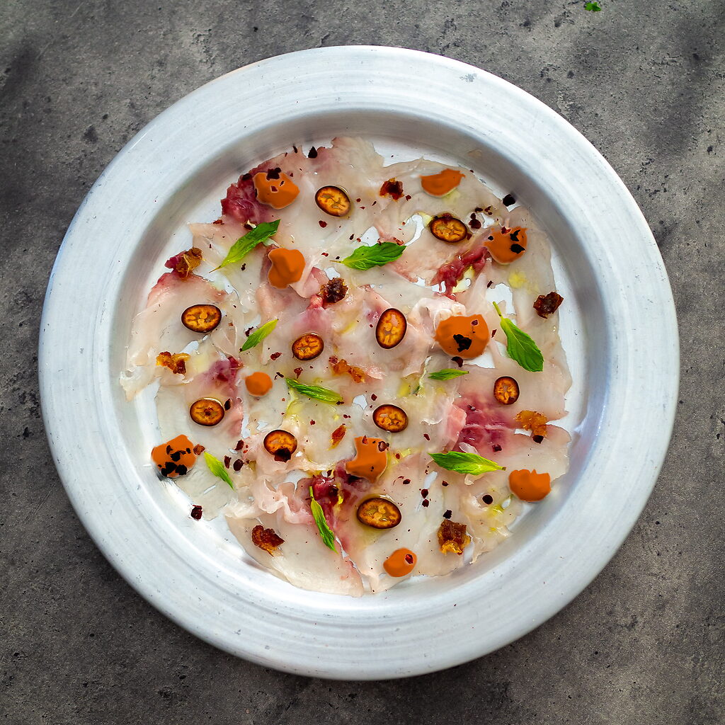 Carpaccio de bar servi sur une assiette avec fines tranches de poisson, agrumes, radis, betterave, grenade et pousses de cresson.