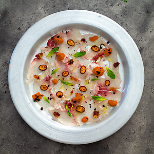 Carpaccio de bar servi sur une assiette avec fines tranches de poisson, agrumes, radis, betterave, grenade et pousses de cresson.